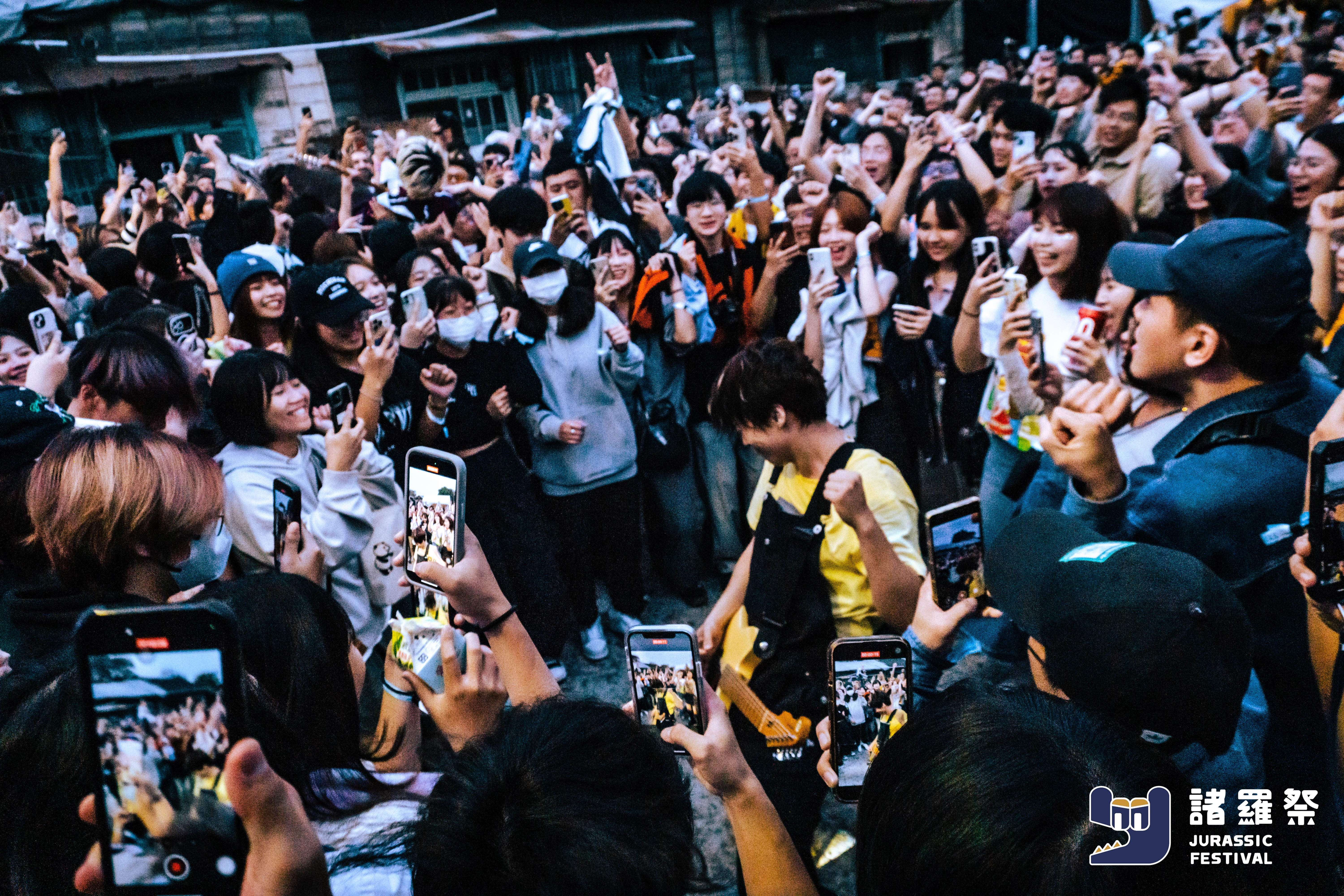 現場觀眾在諸羅祭 Jurassic Festival 熱情圍繞表演者，舉起手機拍攝與歡呼，呈現高度互動的活動體驗氛圍，體現有種創造的活動企劃現場能量。